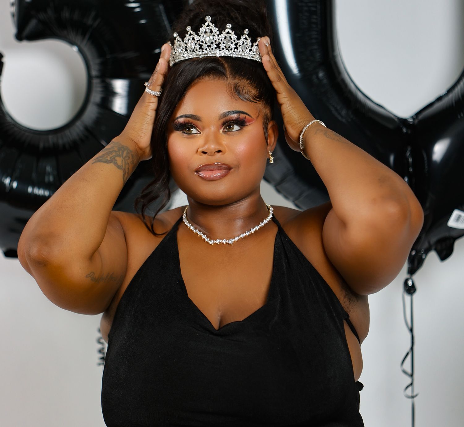Woman wearing crystal tiara and pearl necklace poses with black balloons for milestone birthday celebration.