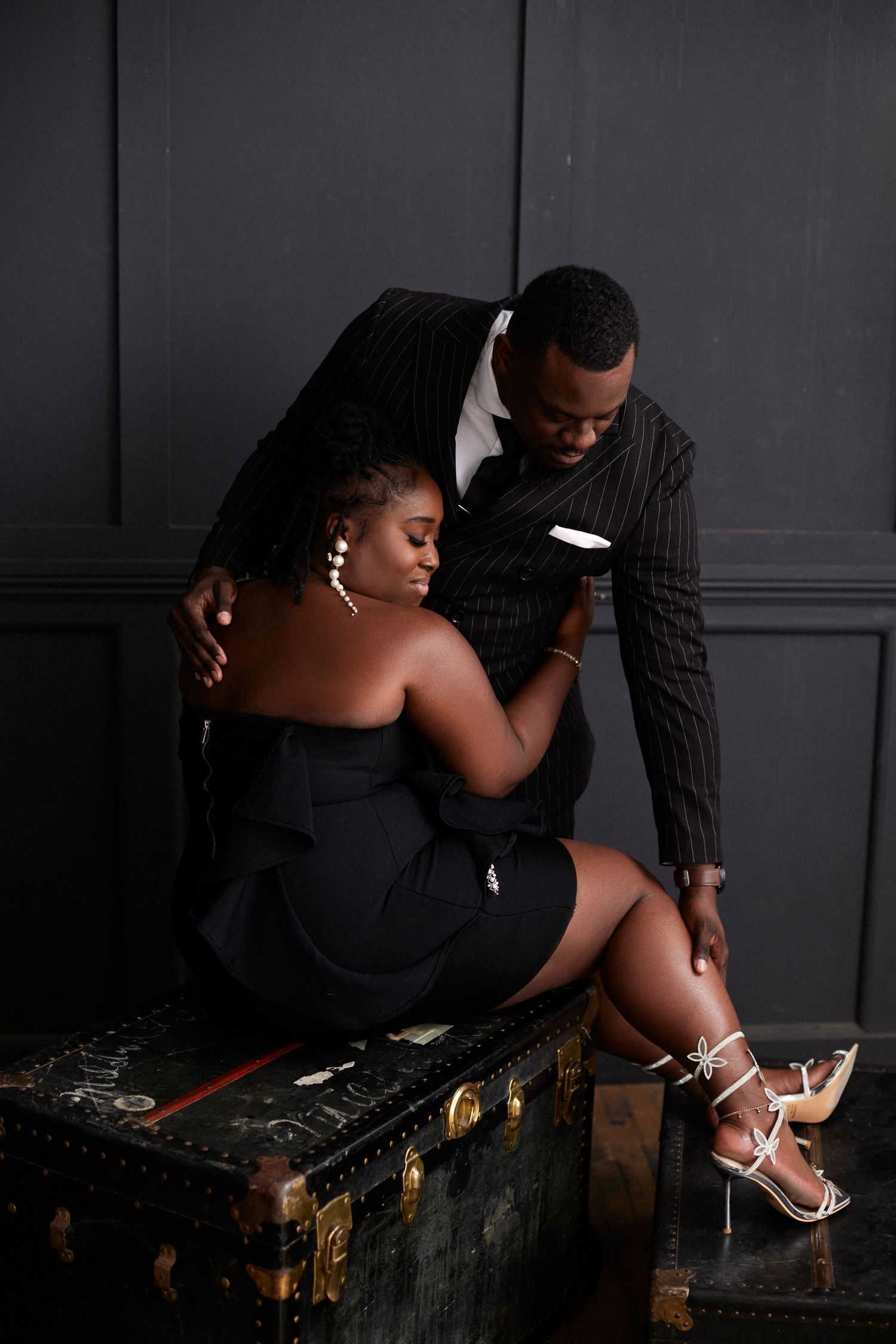 Stylish couple poses on vintage trunk in moody studio setting.