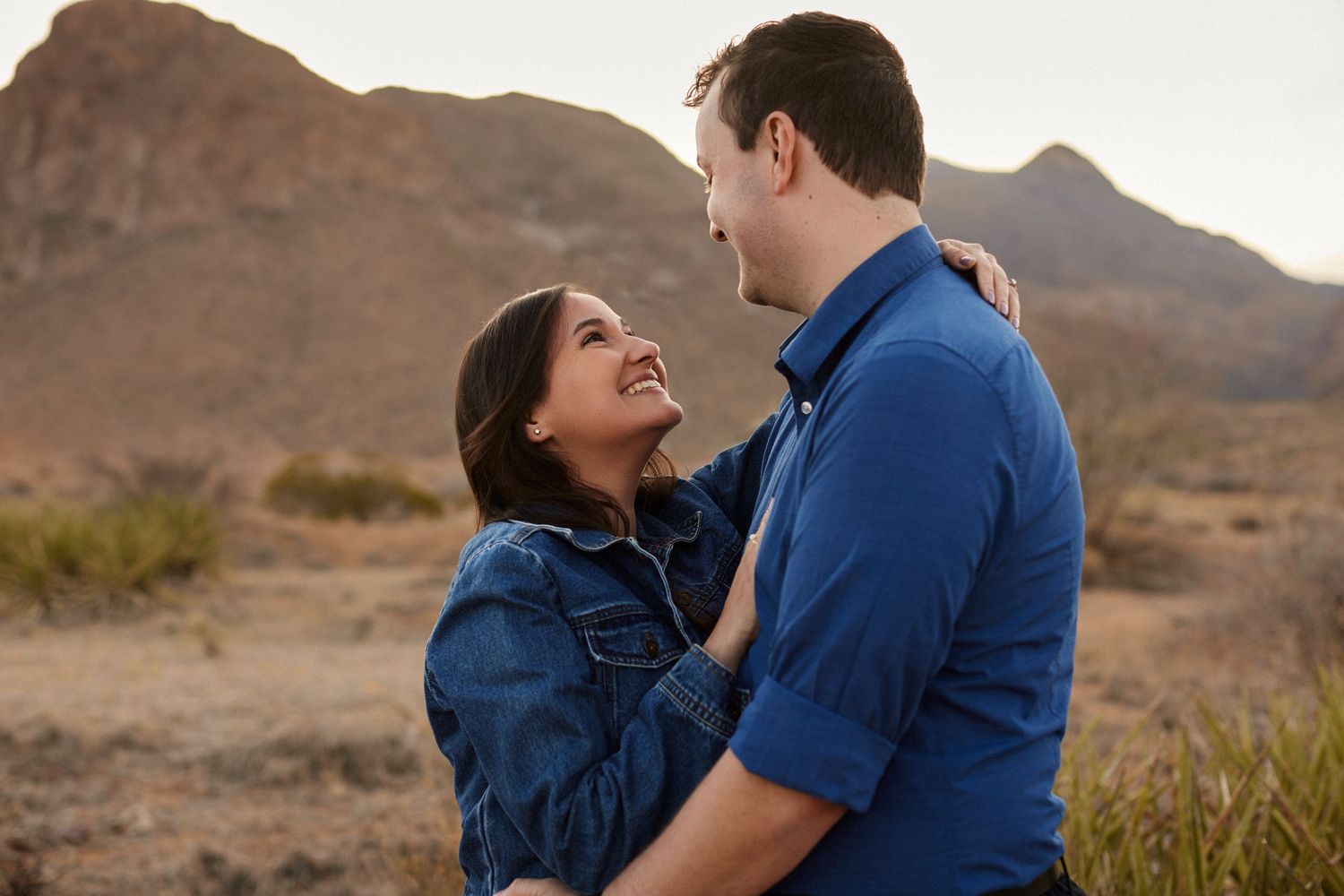 Couple in denim jackets share romantic moment against desert mountain backdrop.
