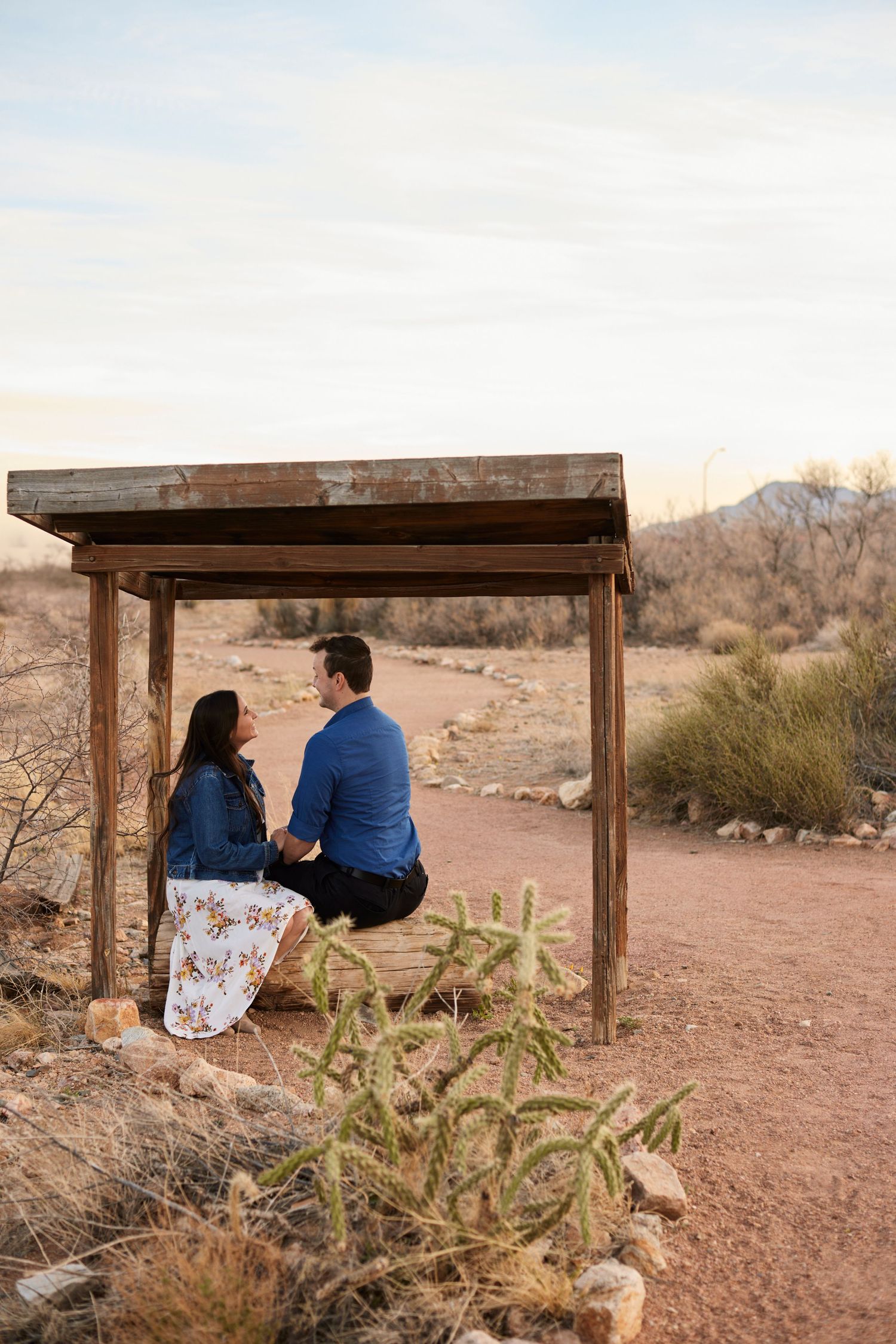 Couple sits together on wooden bench in desert setting with mountains in background.