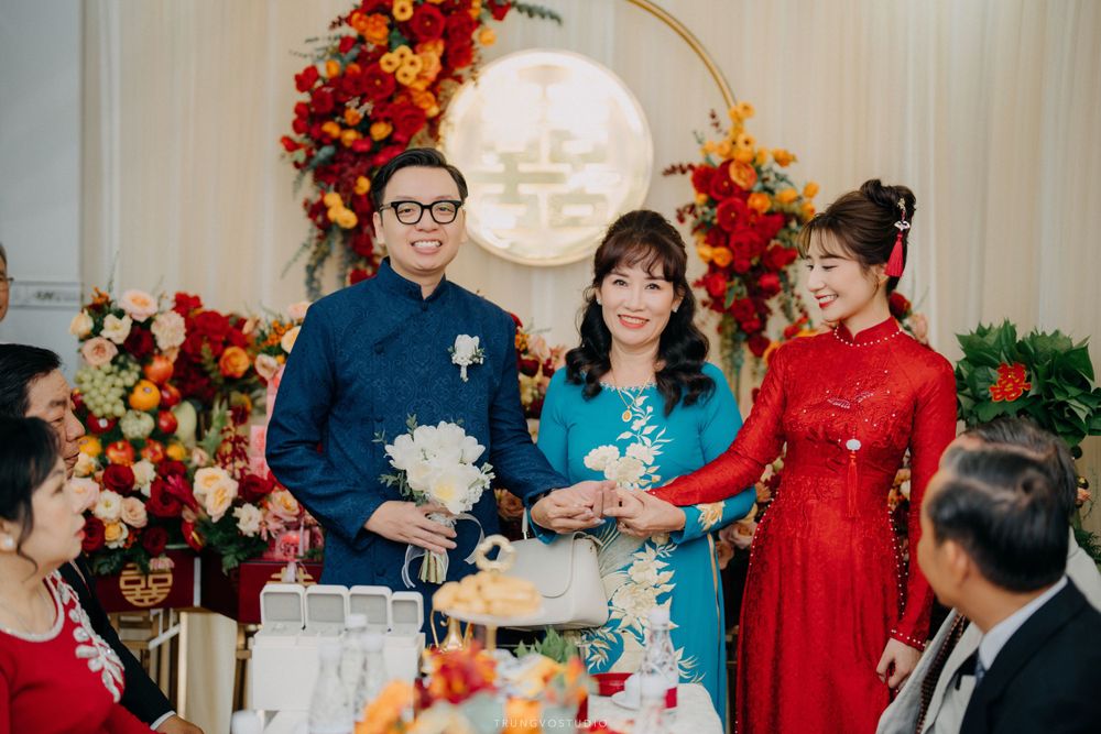 Couple in traditional Vietnamese wedding attire with family at decorated ceremony venue featuring red floral arrangements.