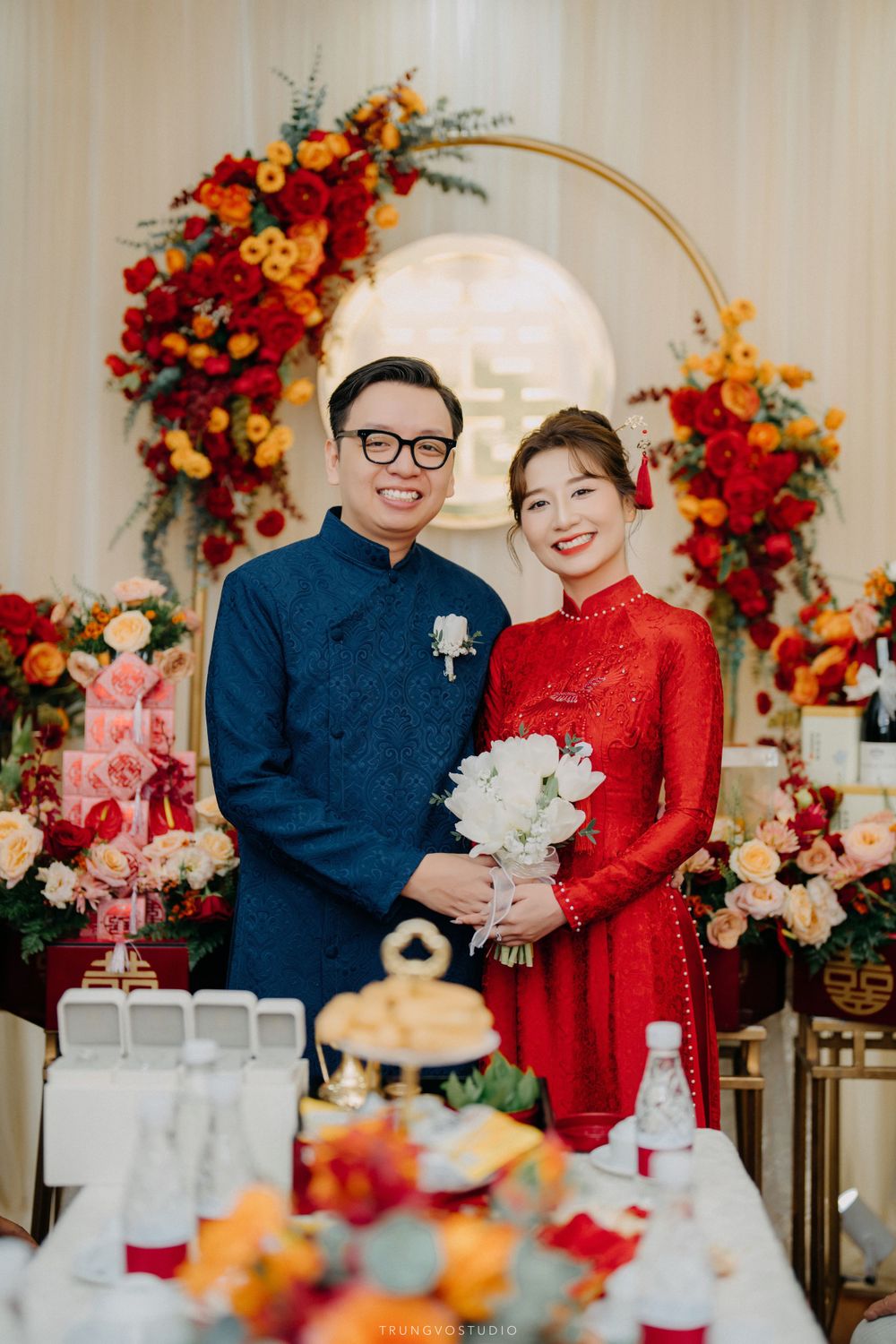 Bride and groom posing in red ao dai and blue ao dai at Vietnamese traditional wedding with floral backdrop.