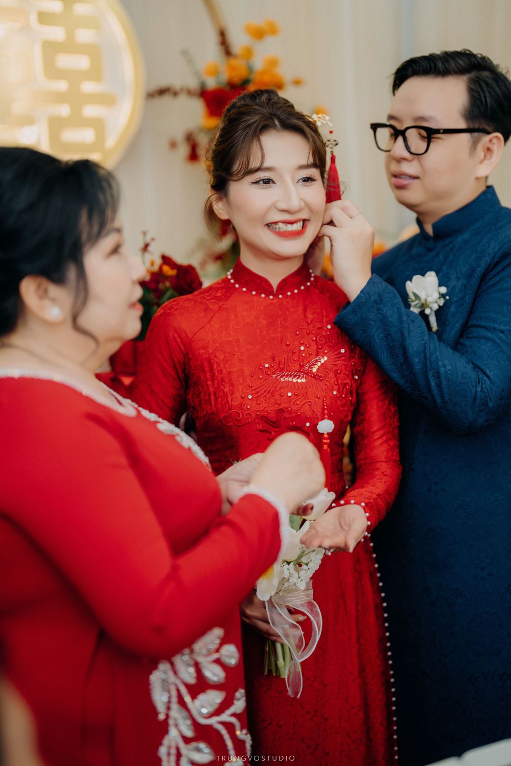 Mother adorning bride with jewelry at Vietnamese traditional wedding ceremony while groom stands nearby.