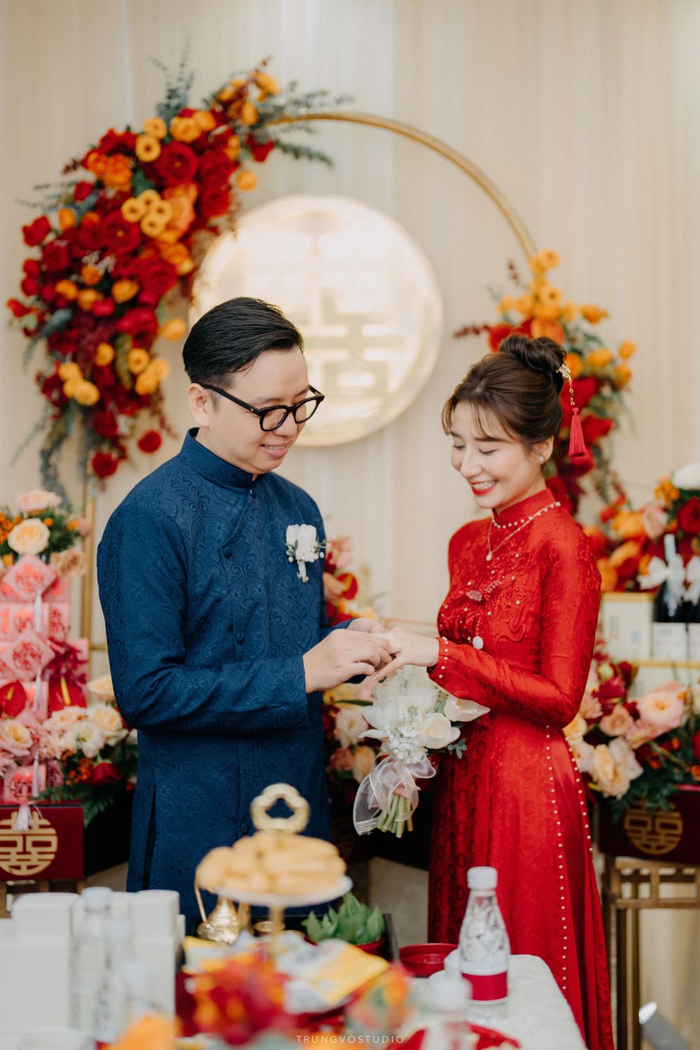 Groom placing ring on bride's finger at Vietnamese traditional wedding with double happiness floral backdrop.