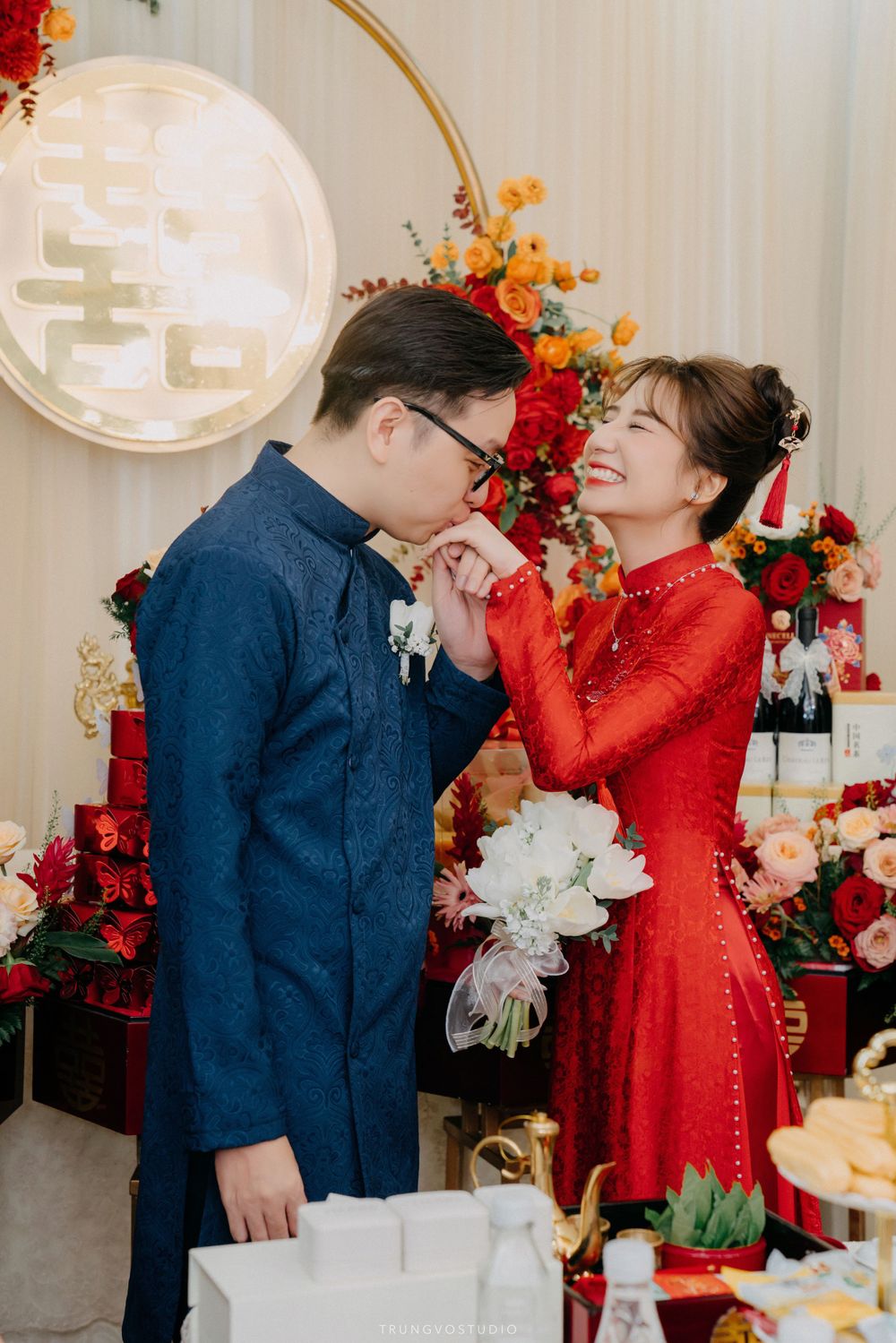 Groom kissing bride's hand at joyful Vietnamese traditional wedding ceremony with floral decorations.