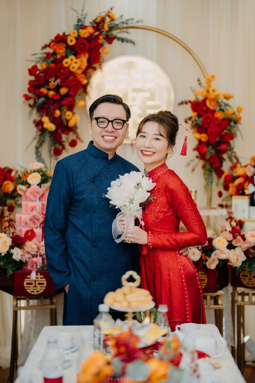 Couple in traditional Vietnamese ao dai attire pose before colorful floral arch at engagement ceremony.