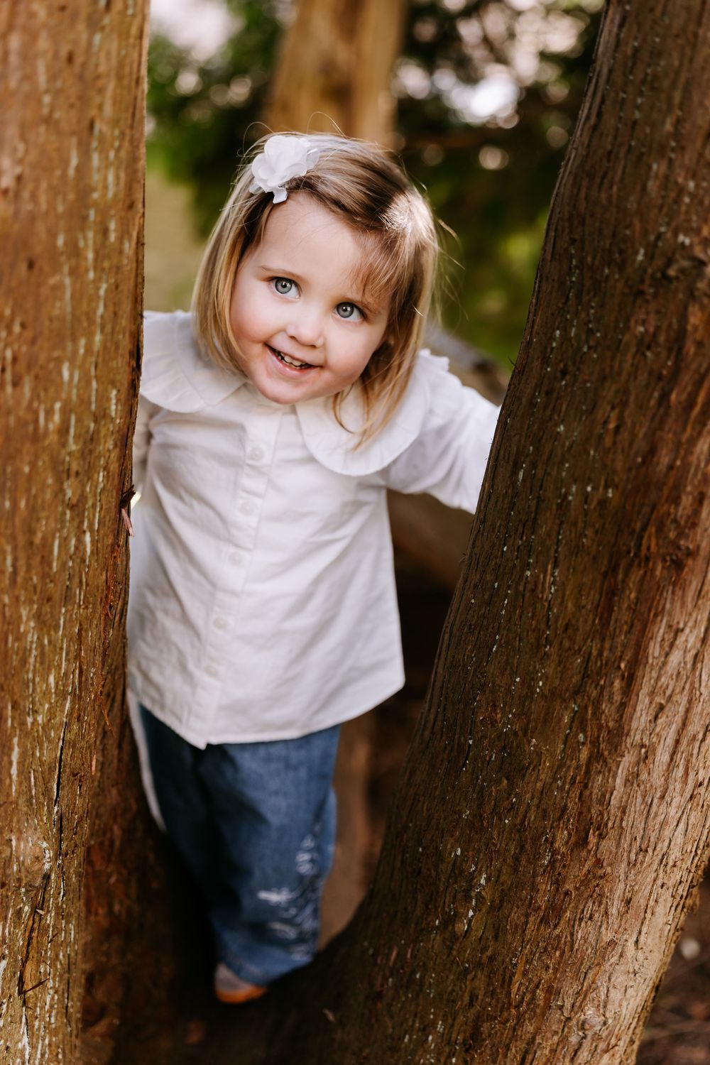 A young child in a white shirt and jeans stands between two tree trunks while smiling at the camera.