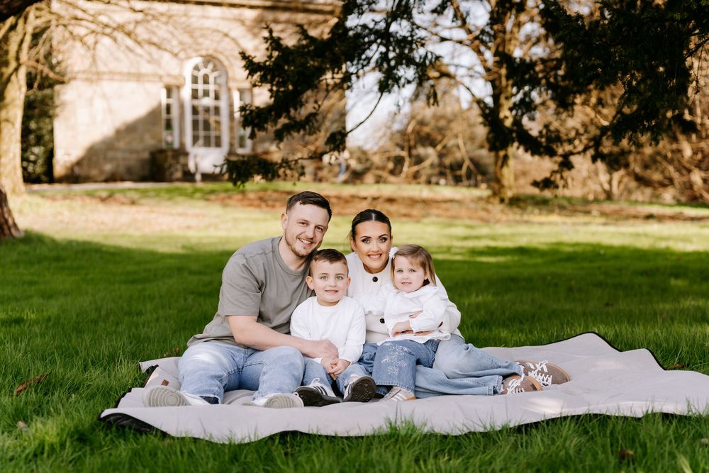 A family sits together on a blanket on a green lawn with a stone building visible in the background.