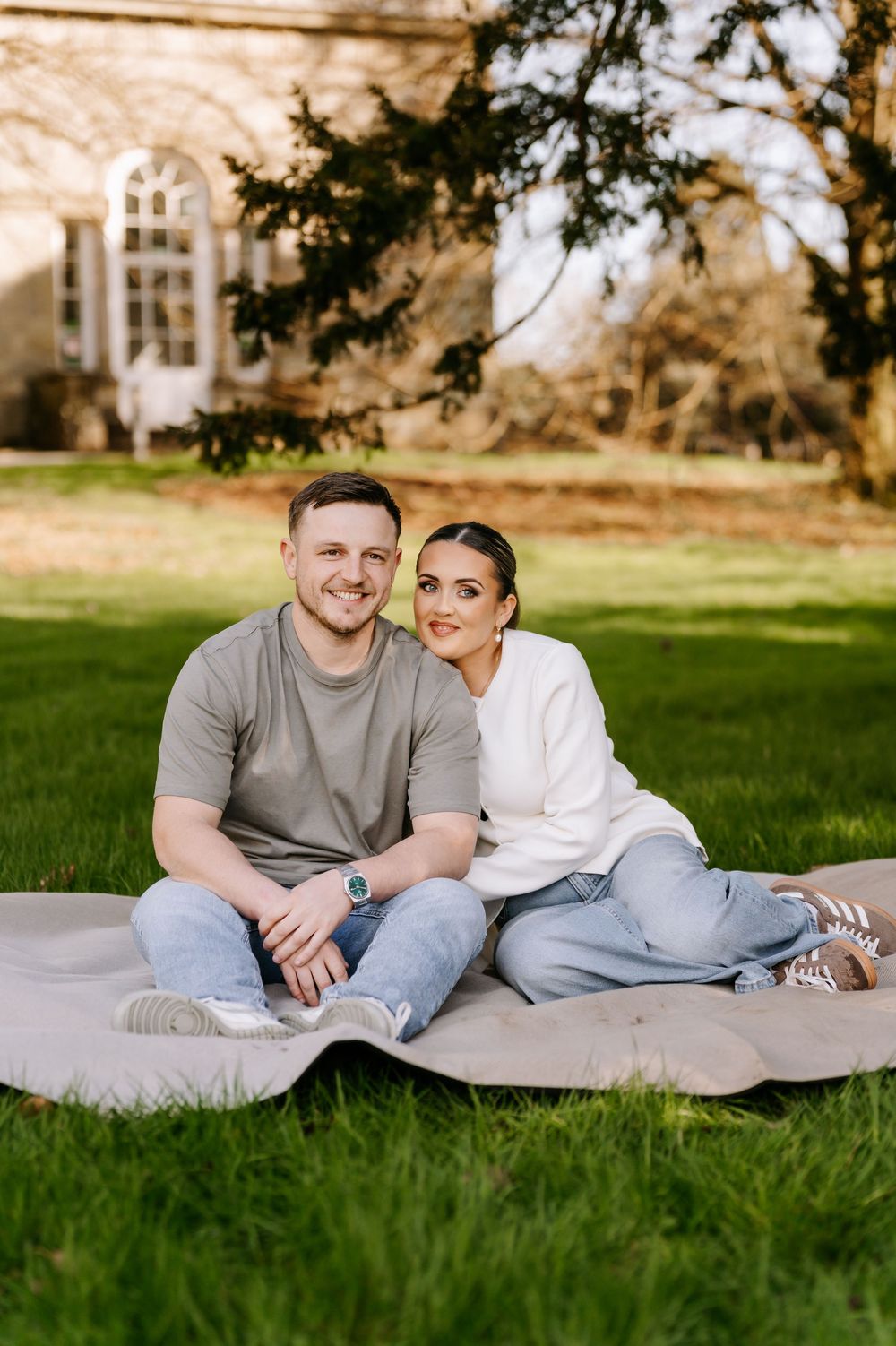 A couple sits together on a blanket on green grass with a building and trees visible in the background.