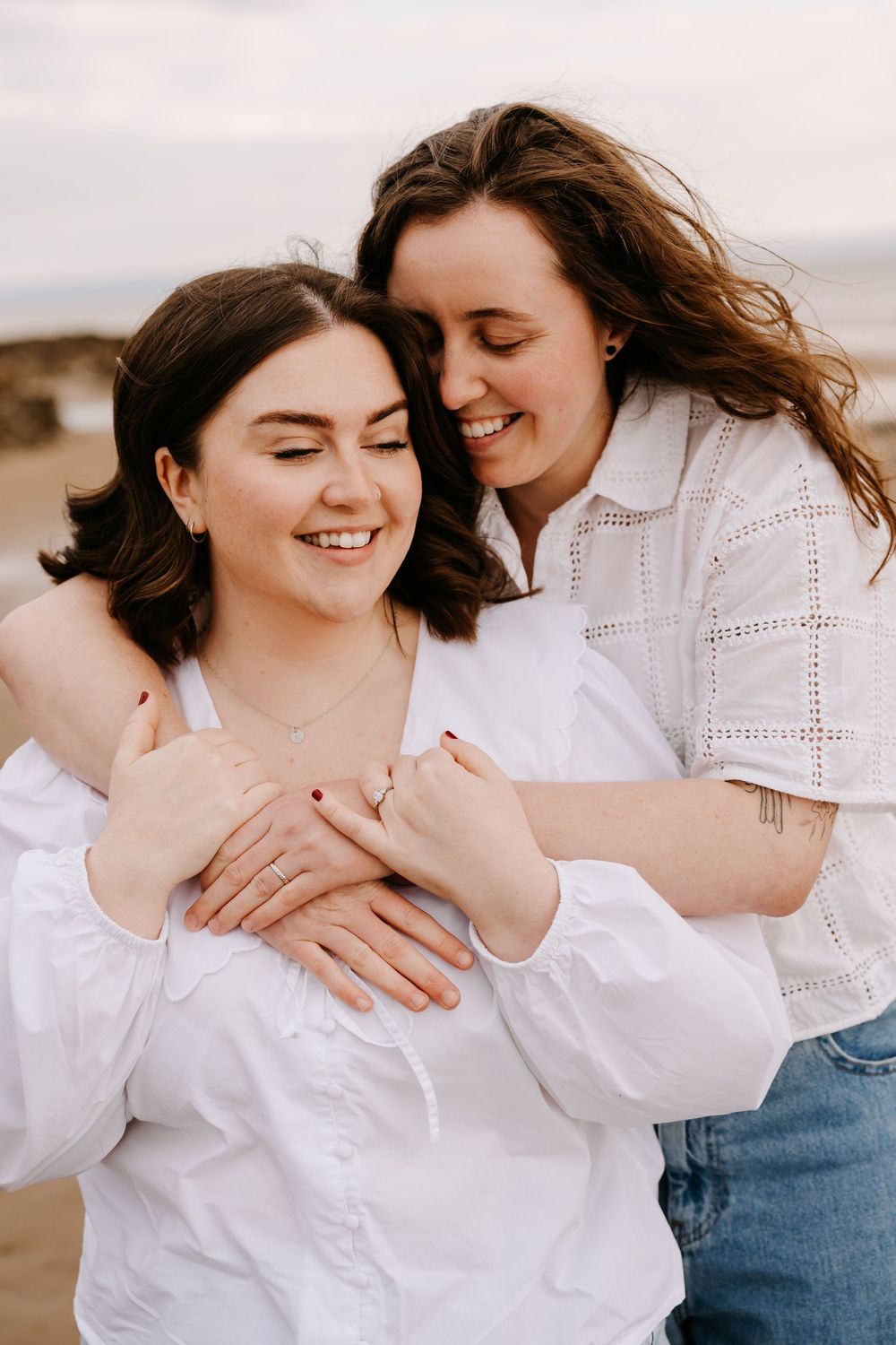 Two women in white tops share a joyful embrace on a beach, one hugging the other from behind while both smile.