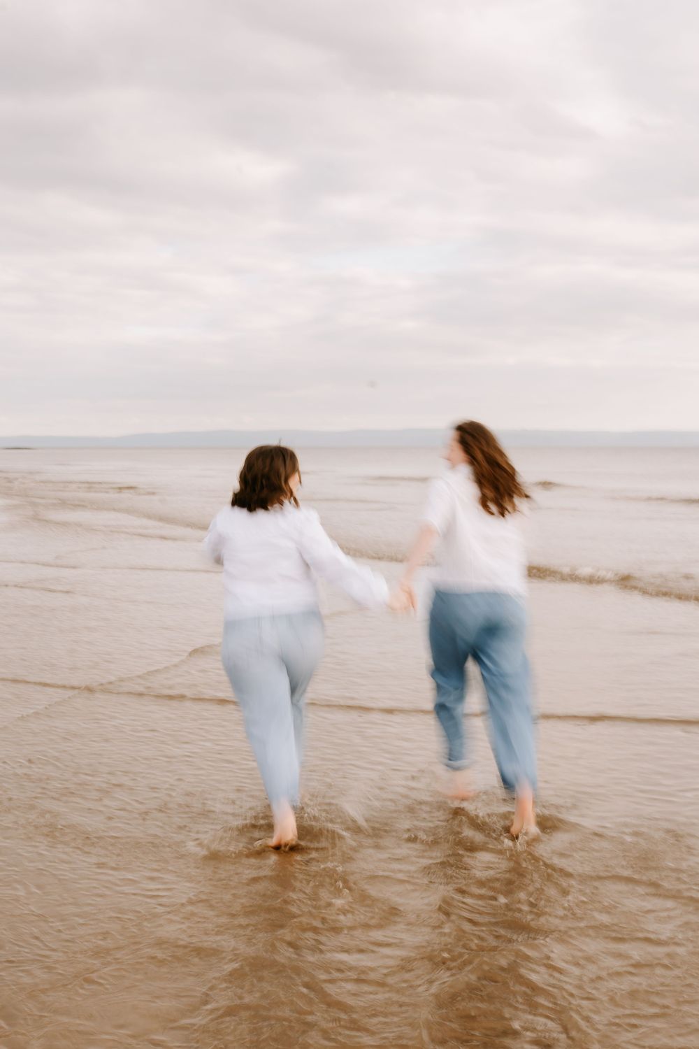 Two women in white tops and blue jeans hold hands running through ocean waves on a cloudy beach day.