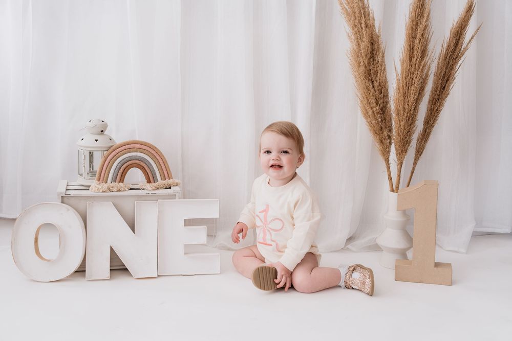 First birthday photography setup with neutral decor featuring wooden 'ONE' letters, pampas grass, and rainbow decorations.