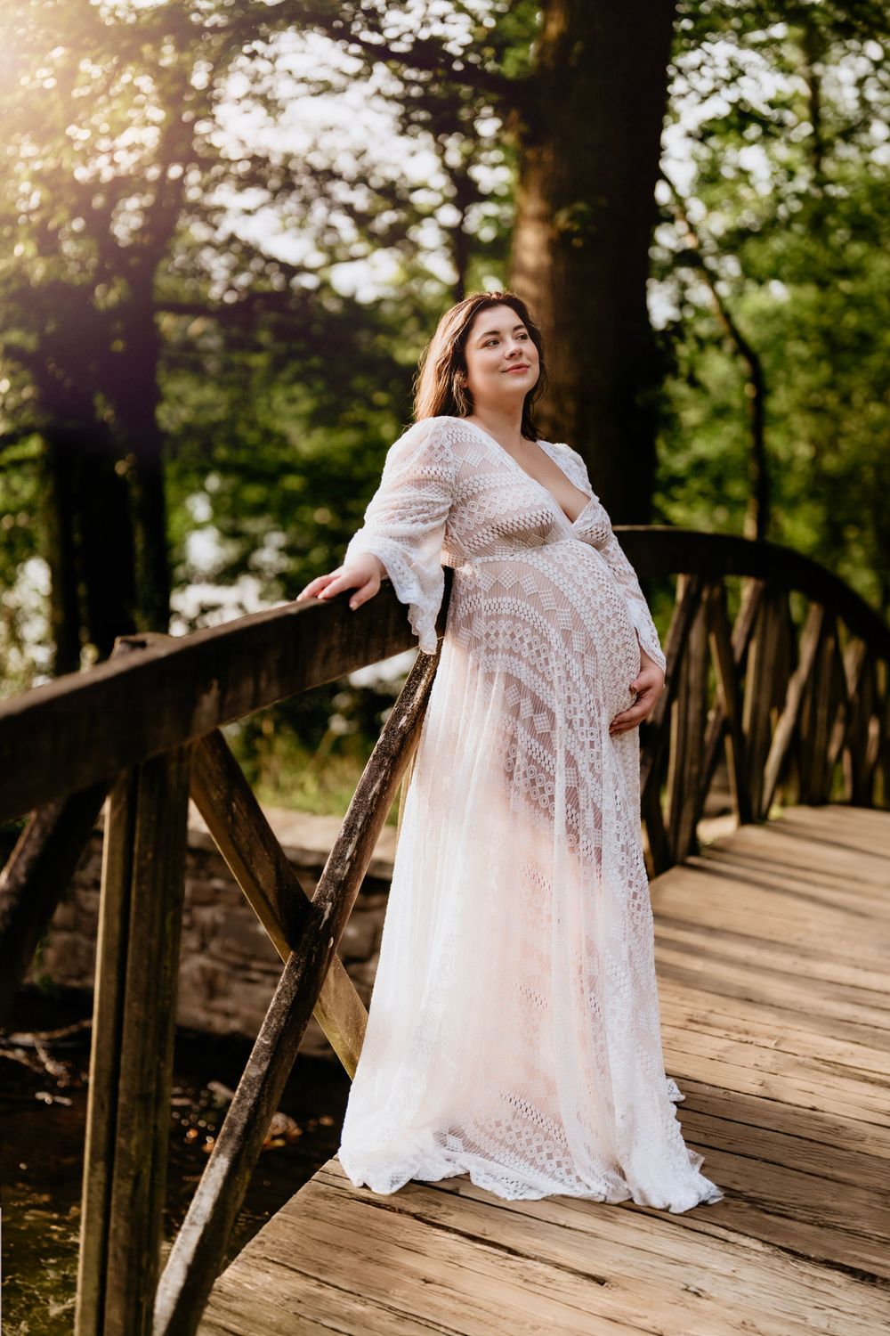 A pregnant woman in a flowing white lace dress poses on a wooden bridge in a sunlit forest setting.