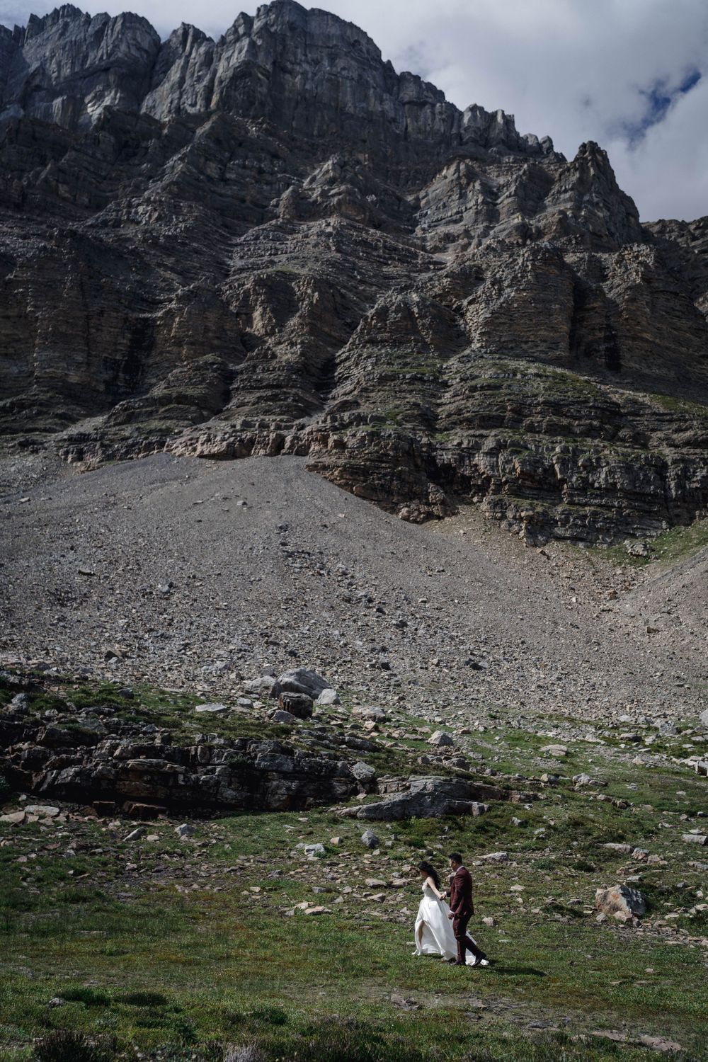 Two figures in wedding attire stand amid a majestic rocky mountain landscape with dramatic cliffs towering above them.