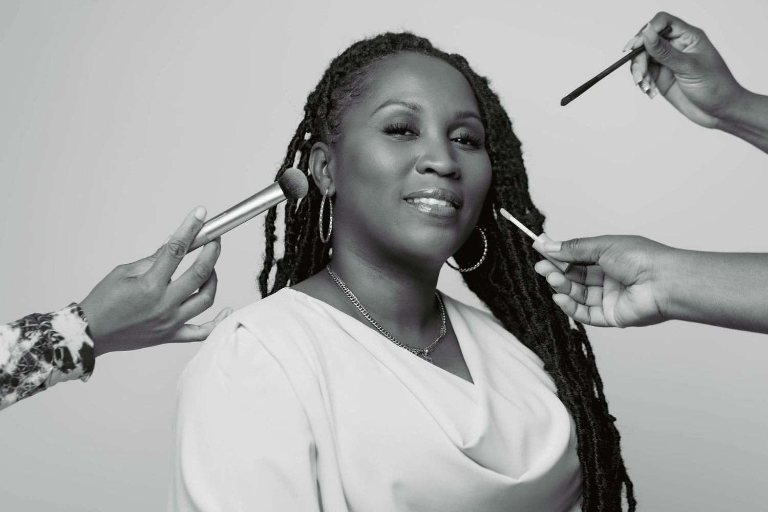 Black and white photo of a woman with long braids getting makeup applied by two artists on a gray background.