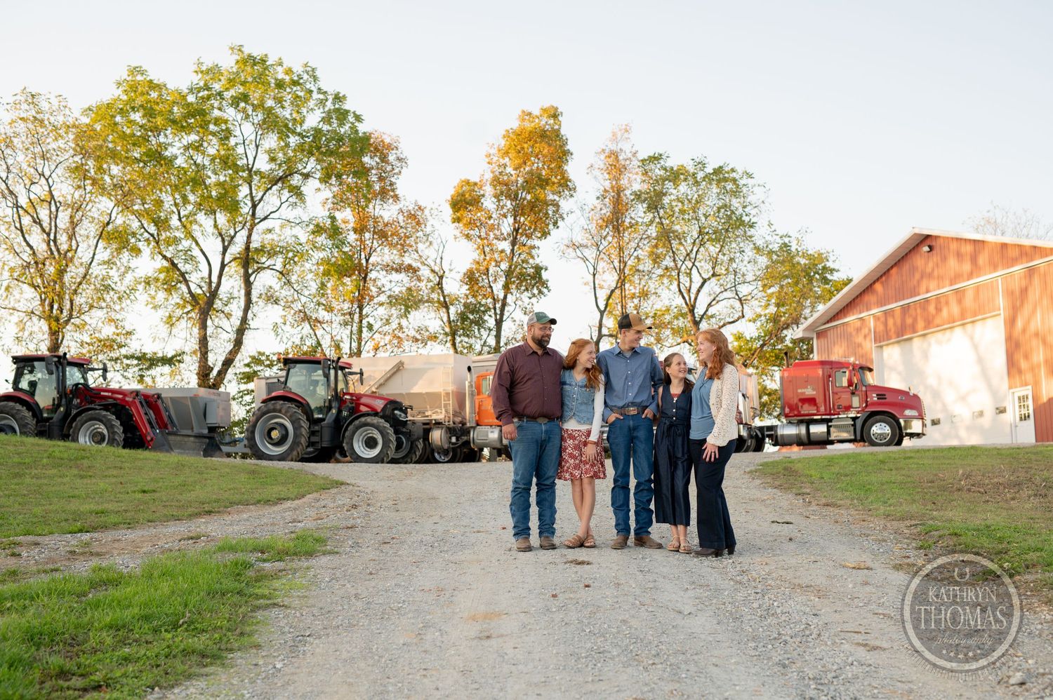 Family poses on gravel path with tractors and farm equipment in autumn setting.