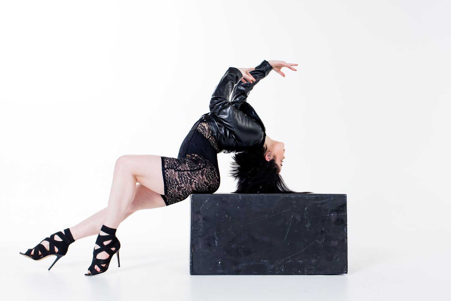 A performer in black leather and high heels poses artistically on a dark cube platform.