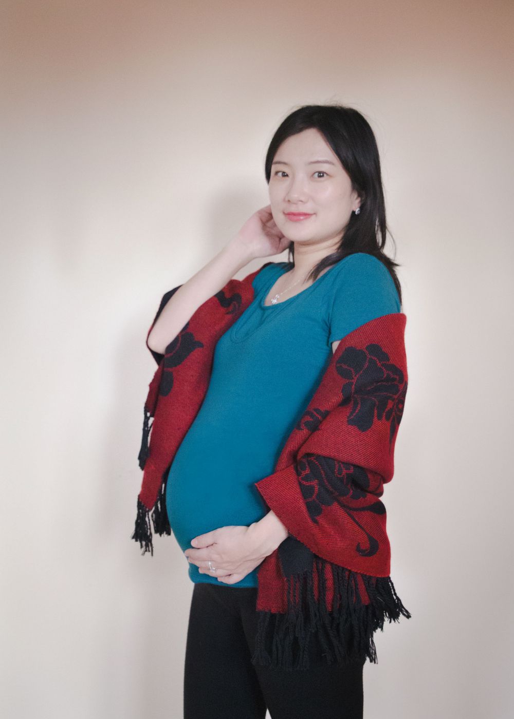 Pregnant woman in teal top and red patterned shawl posing against light background.