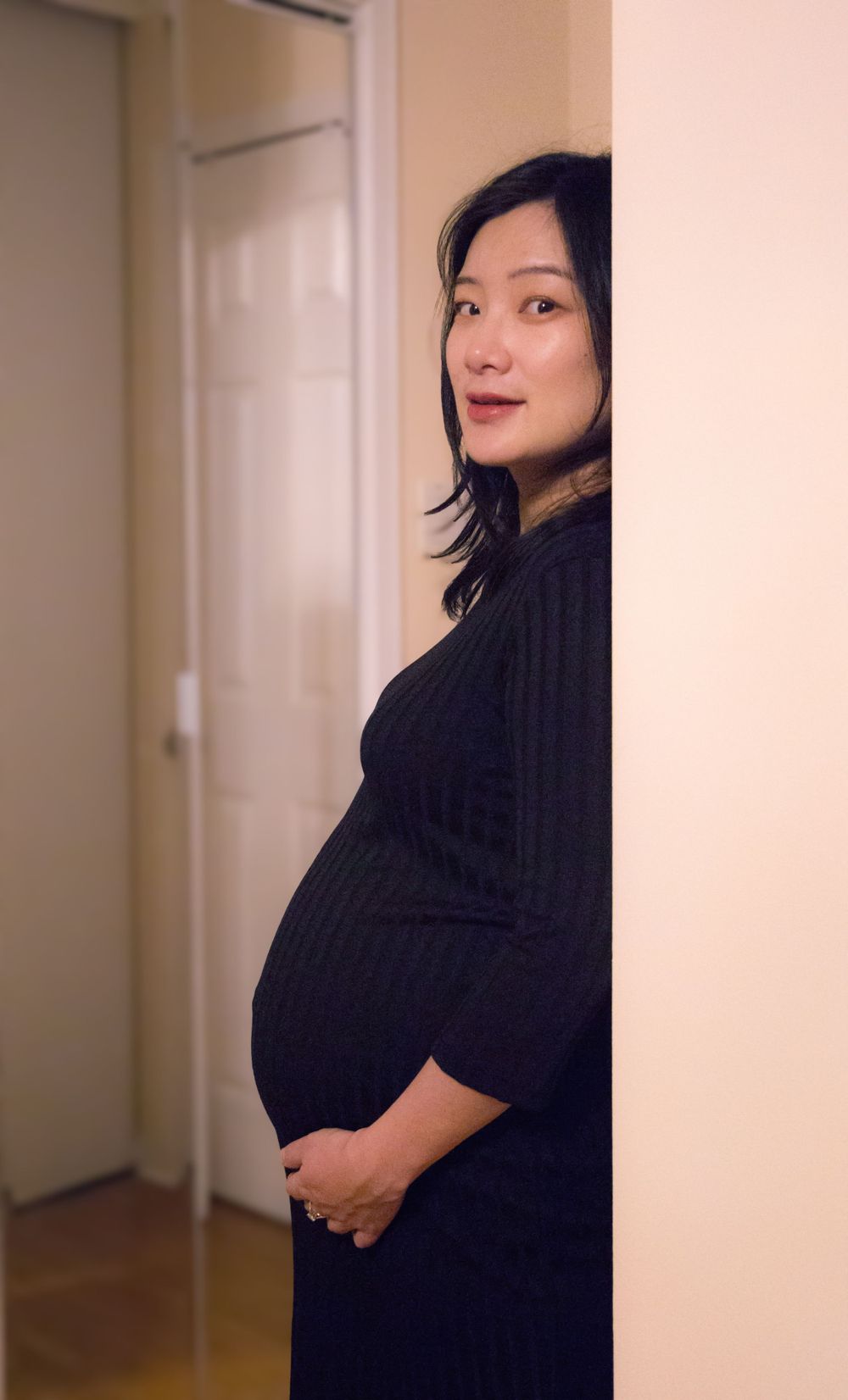 Artistic maternity portrait of woman in black dress peeking from behind wall.