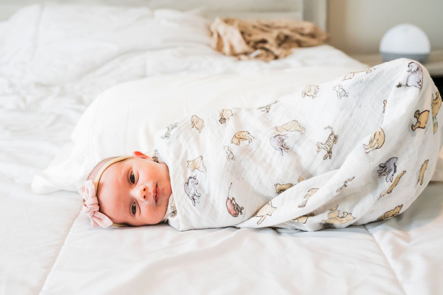 A baby wrapped in a white floral swaddle blanket lies on white bedding.