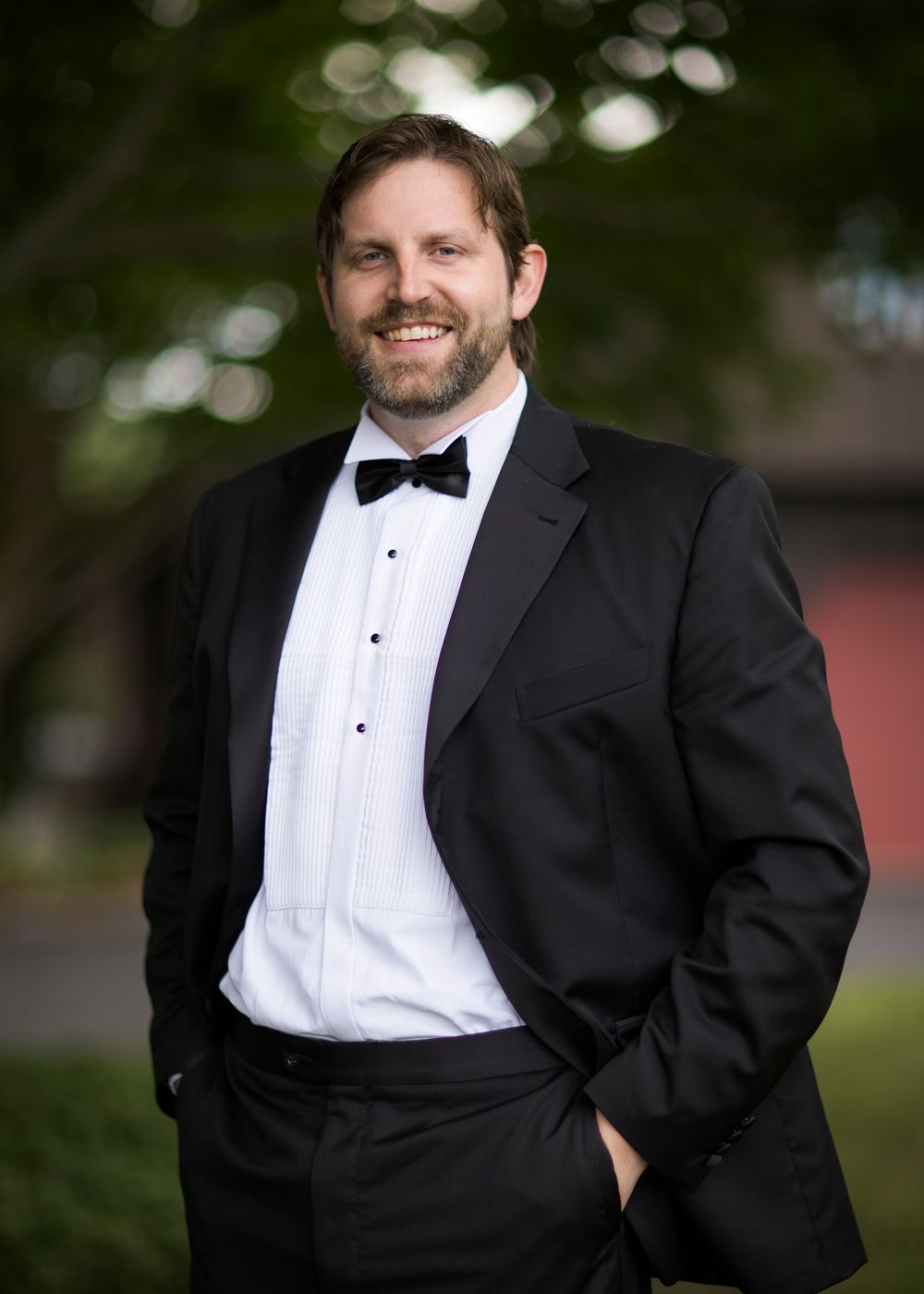 A groom in a classic black tuxedo with bow tie poses outdoors for formal wedding portraits.