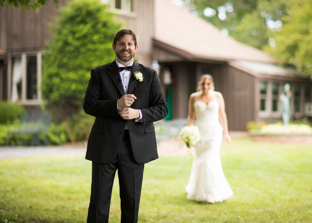 A wedding couple shares a playful moment on a green lawn with the groom in the foreground and bride in the distance.