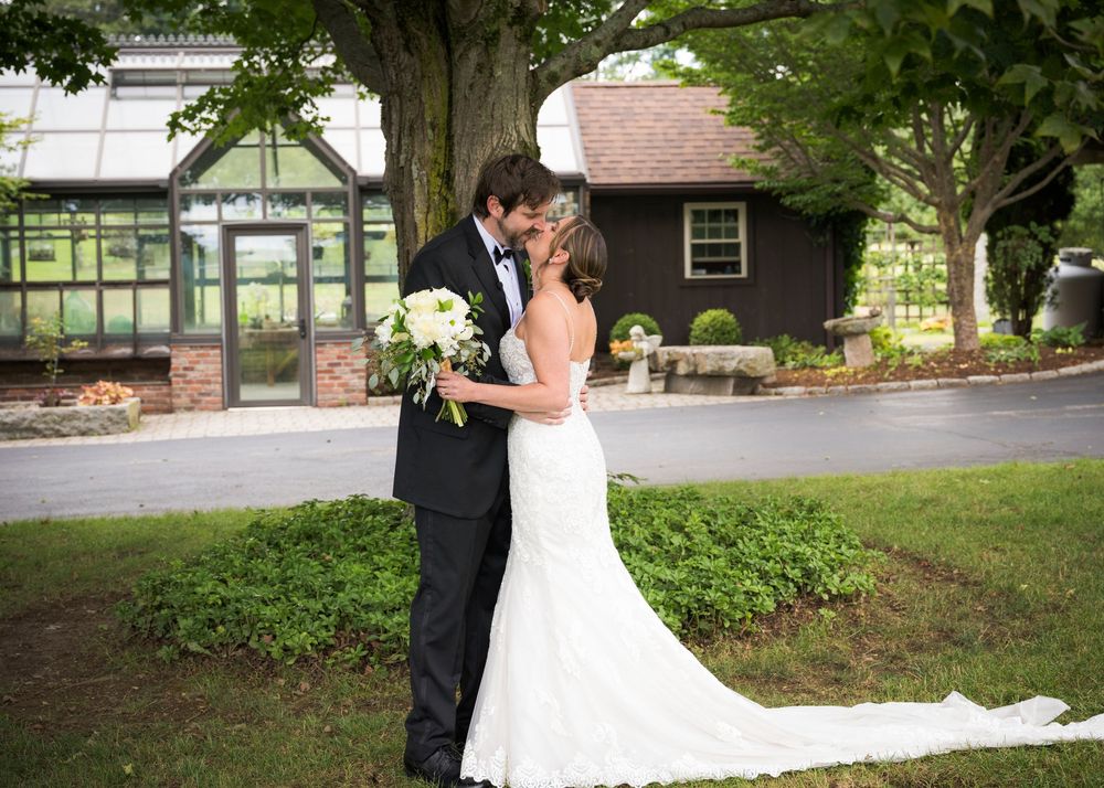 A romantic wedding moment captured under a large tree with lush greenery and a scenic pathway in the background.