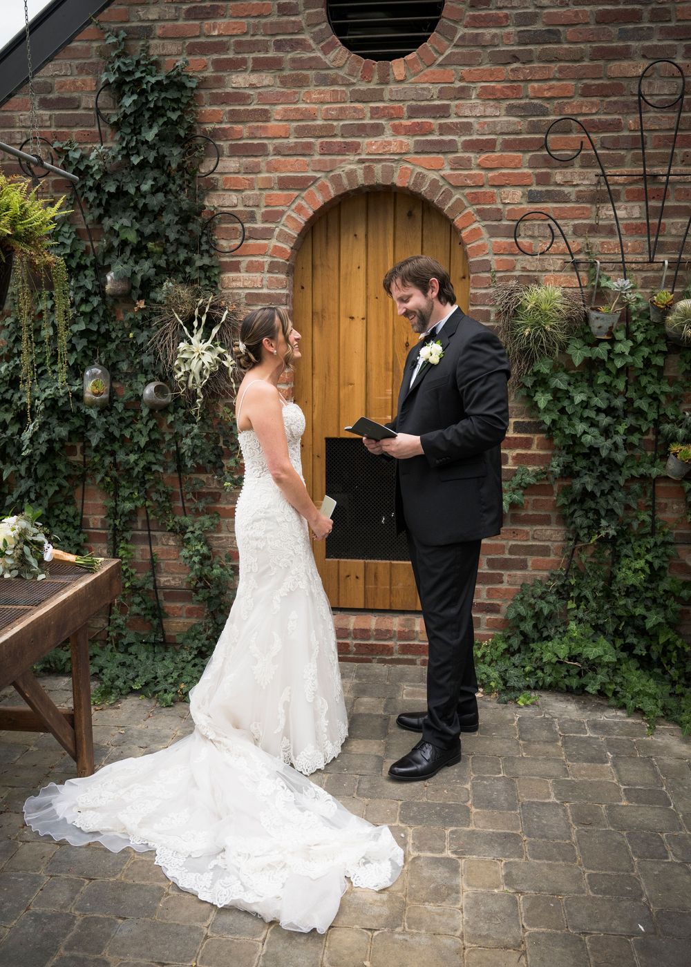 A wedding couple shares a moment in front of a rustic brick wall and wooden door adorned with climbing ivy.