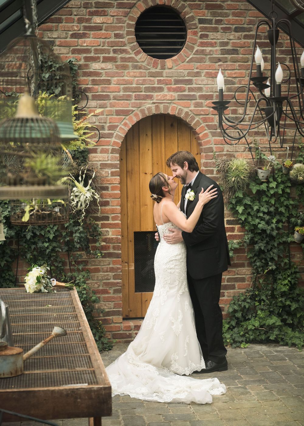 A romantic wedding couple embraces in front of a rustic brick building with wooden doors and climbing vines.