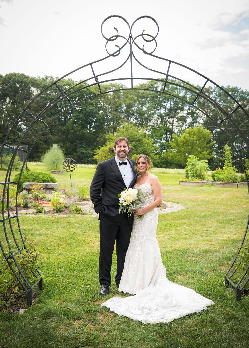Wedding couple stands beneath an ornate metal garden arch on a lush green lawn during their outdoor ceremony.