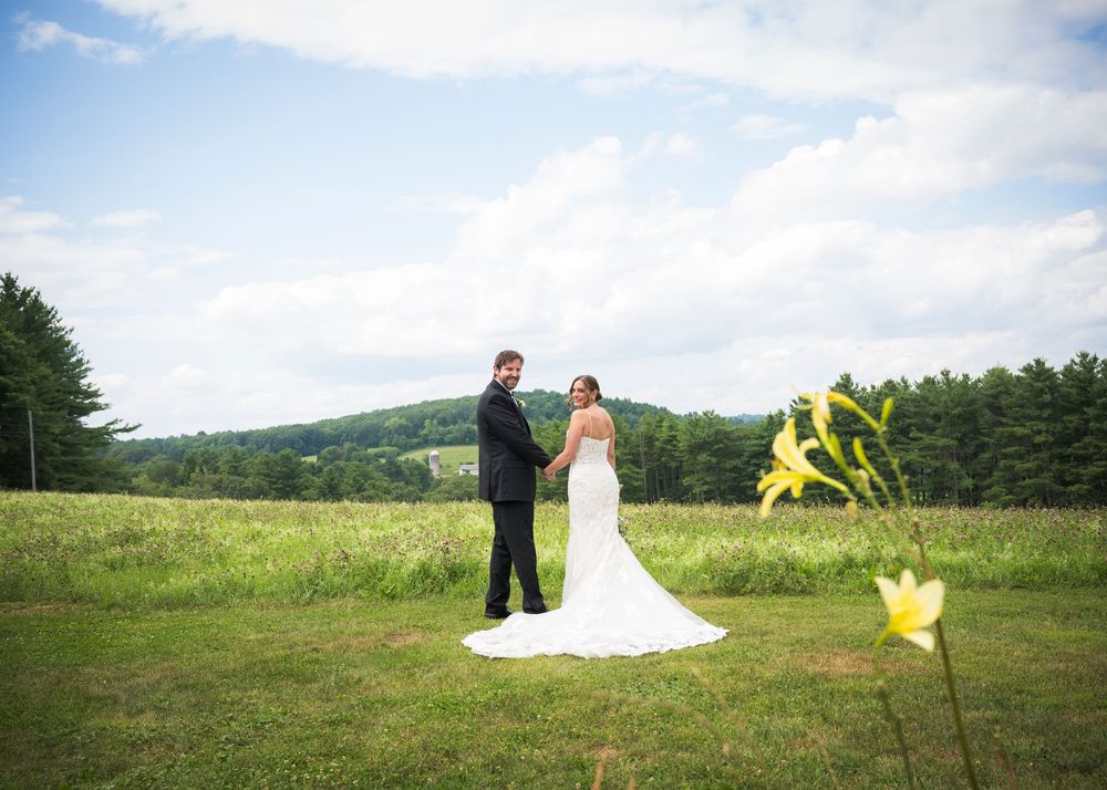 A wedding couple stands in a scenic meadow with rolling hills and yellow wildflowers on a bright summer day.