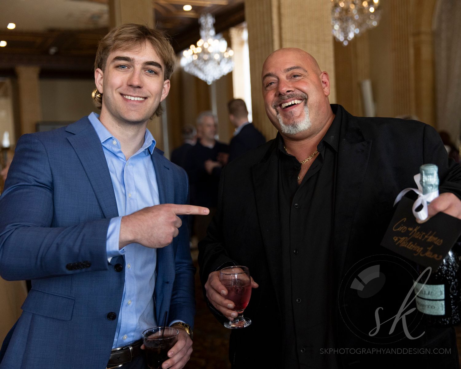 Two men smiling and posing at an elegant event, one in a blue suit pointing, the other in black holding a drink.