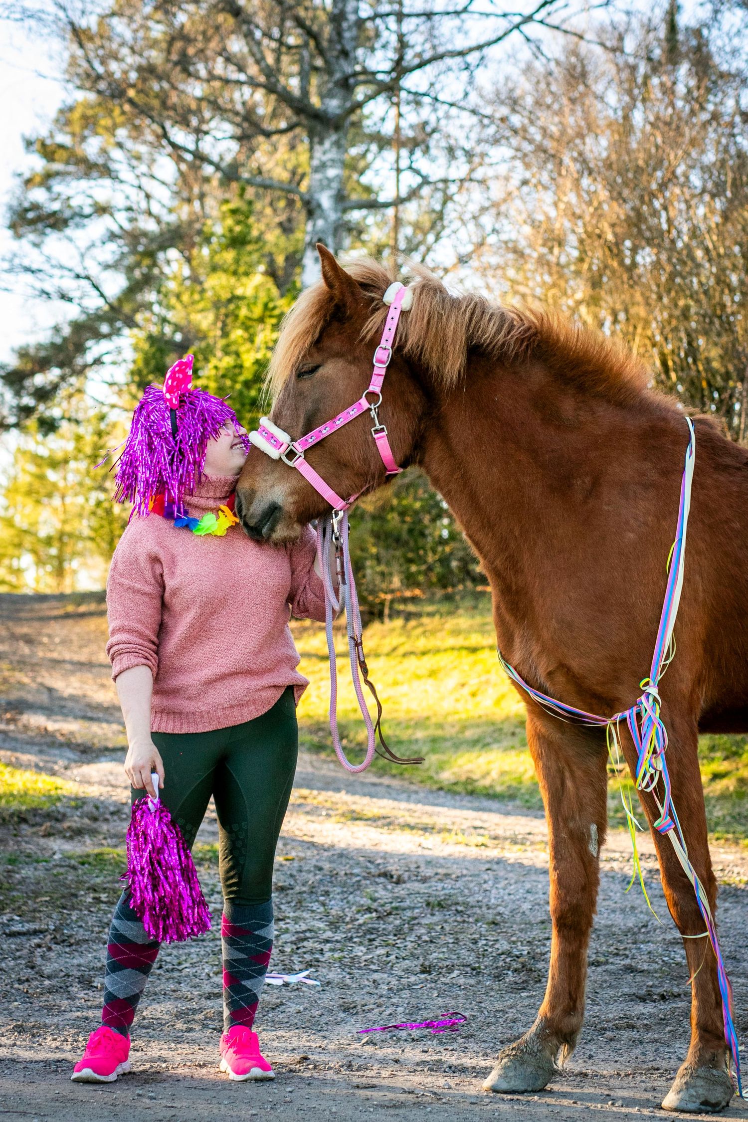 Someone wearing a bright purple wig and pink sweater stands next to a brown horse wearing pink tack outdoors.