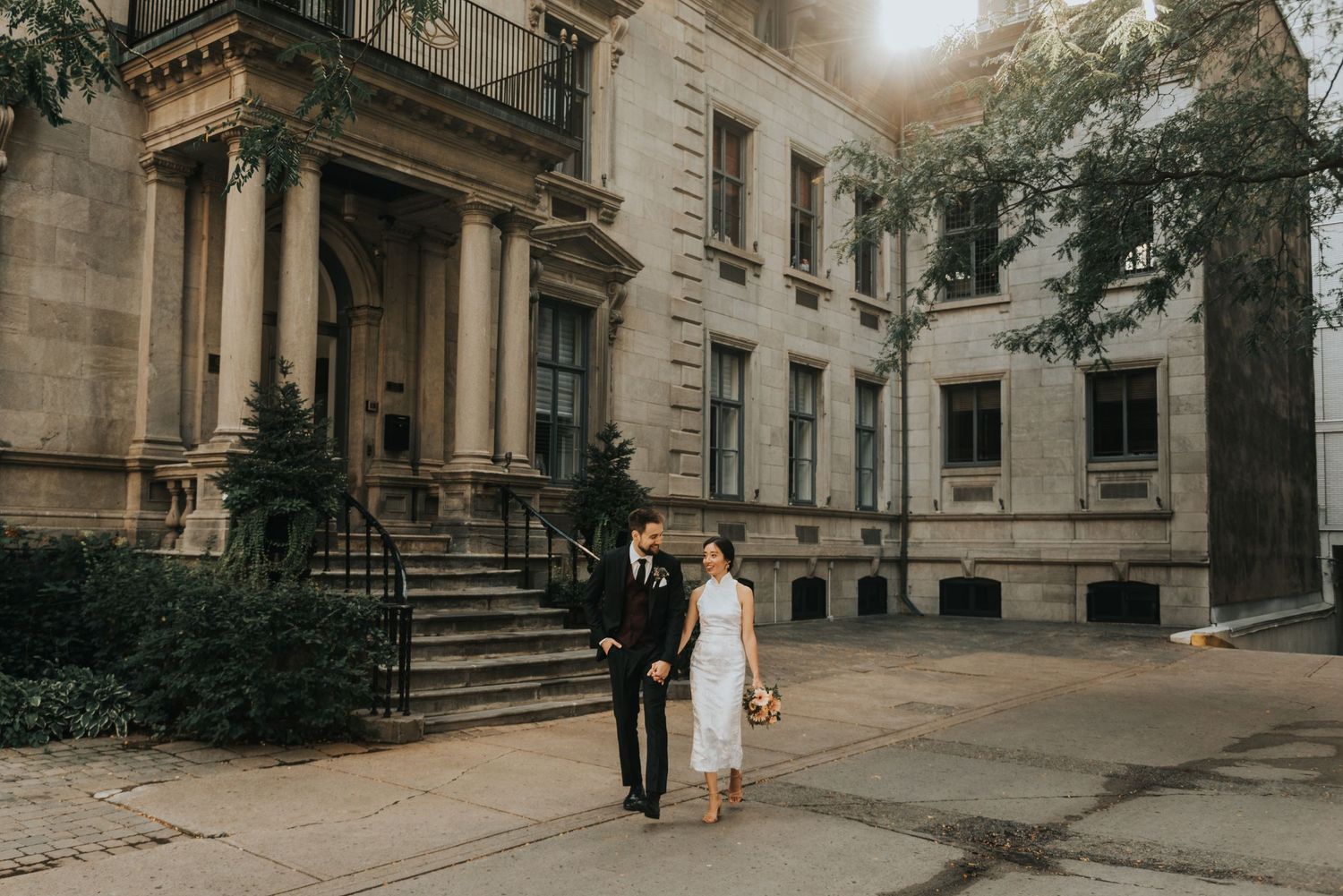 A couple stands together outside a grand historic mansion with stone columns and elegant architecture on an overcast day.
