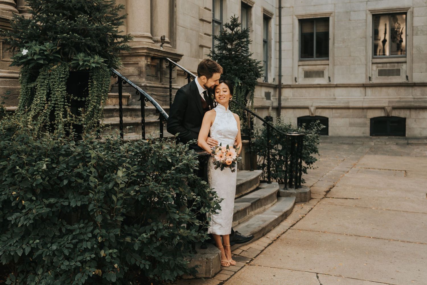 A romantic couple embraces on the stone steps of a historic building with green foliage nearby.