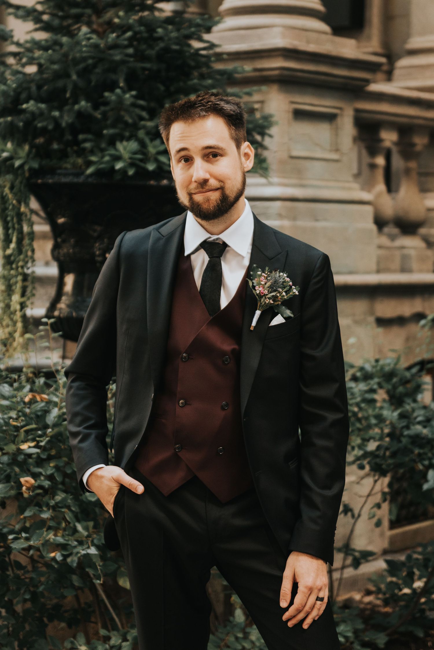 A groom wearing a black suit with burgundy vest and boutonniere stands in front of stone architecture and greenery.
