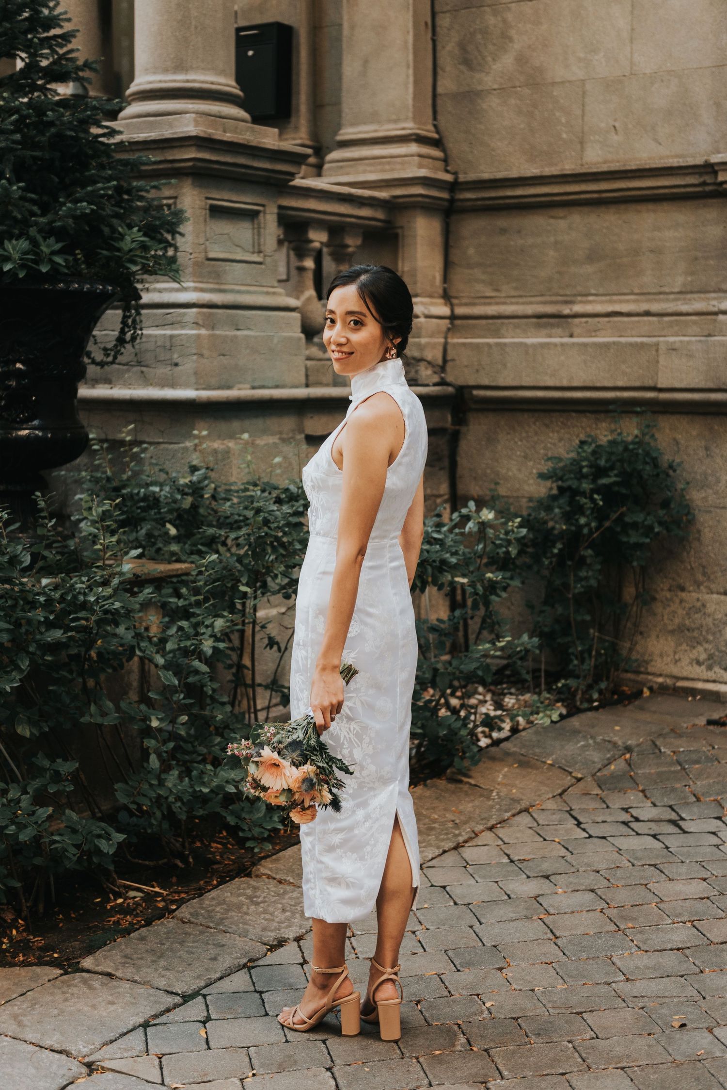 A white lace qipao-style dress worn at an elegant stone building with columns and cobblestone path.