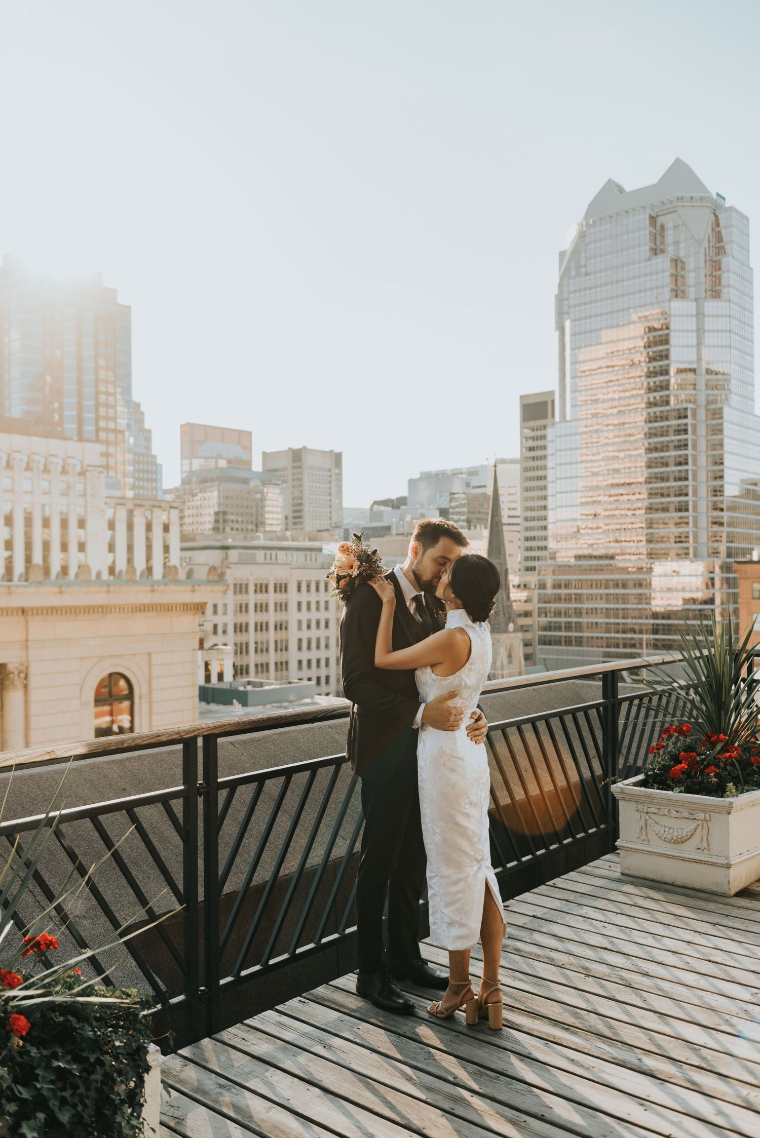 A romantic couple embraces on an urban rooftop terrace with modern skyscrapers and city buildings in the background.