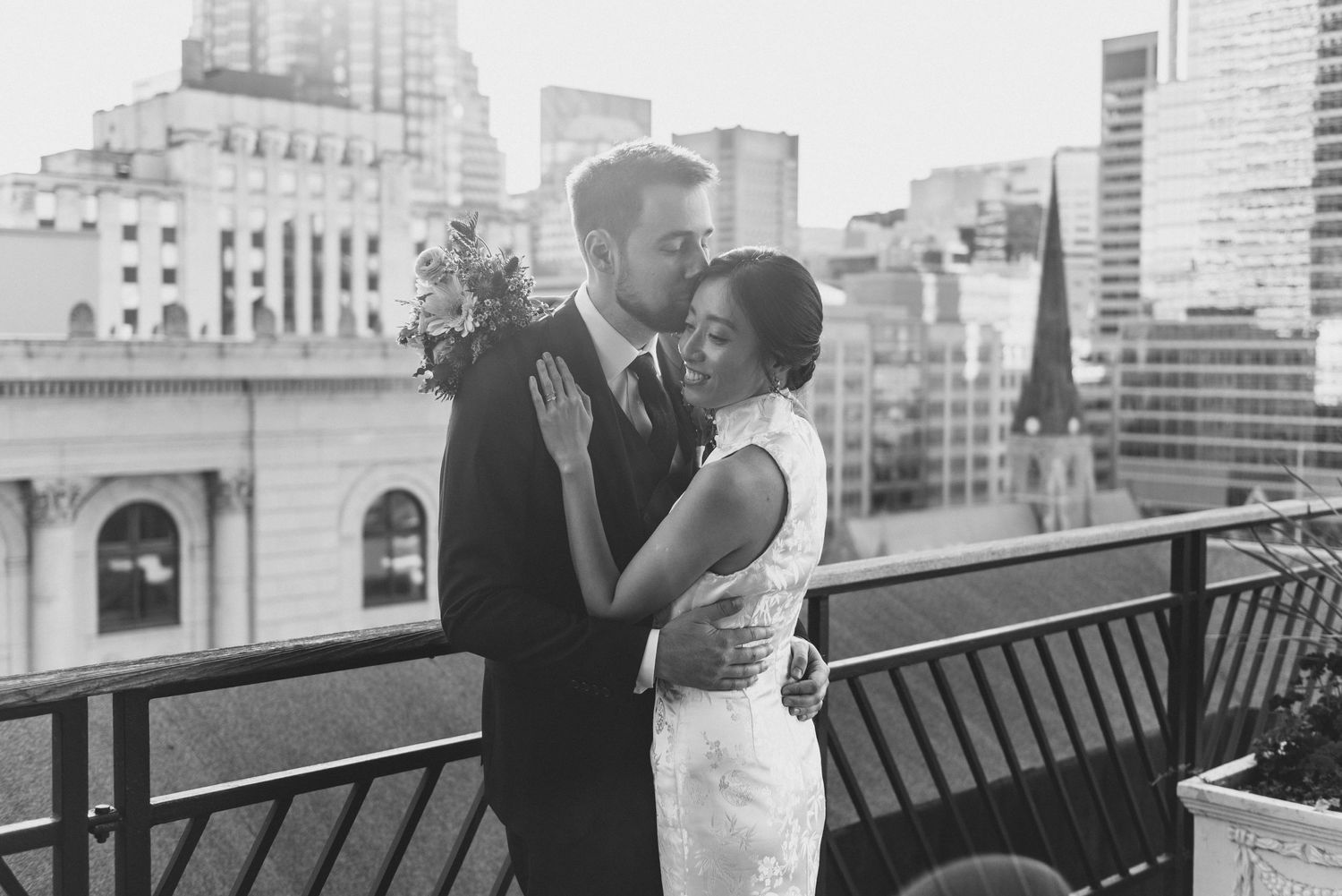 A romantic couple shares an intimate moment on an urban rooftop terrace with city buildings in the background.