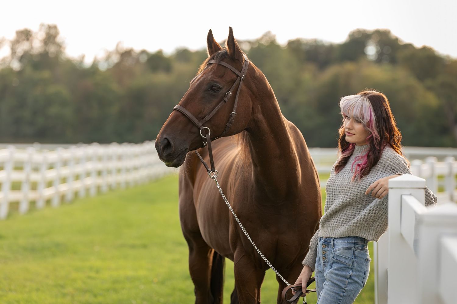 A young woman with pink hair stands beside a chestnut horse at a white fence ranch on a sunny day.