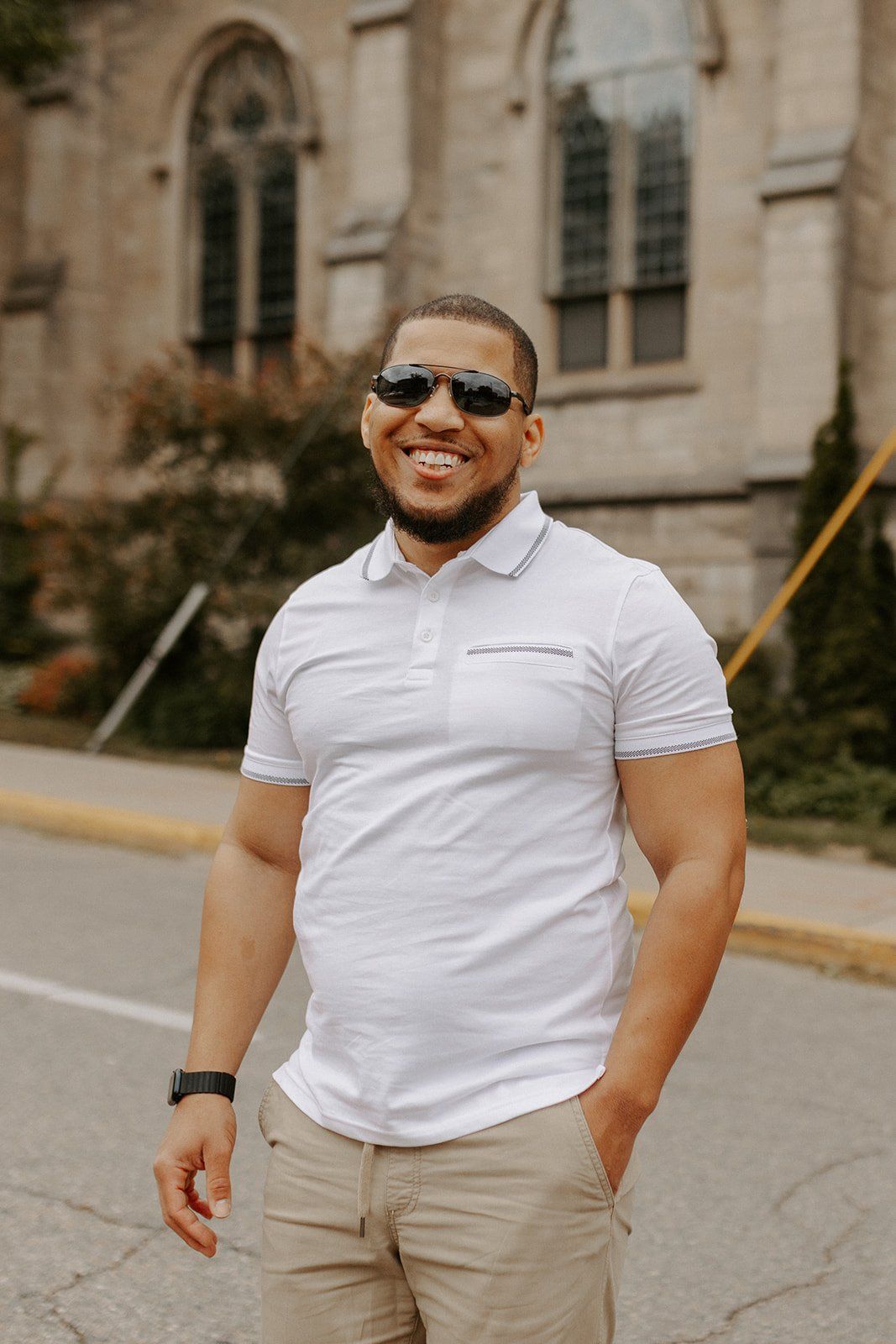 A person wearing a white polo shirt and khaki pants stands confidently outdoors near a stone building.