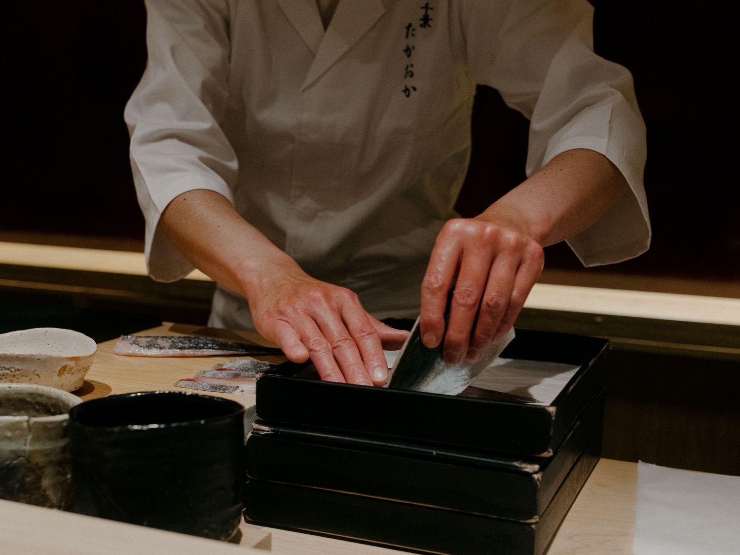 Hands preparing sushi at a traditional wooden counter with black preparation surface.