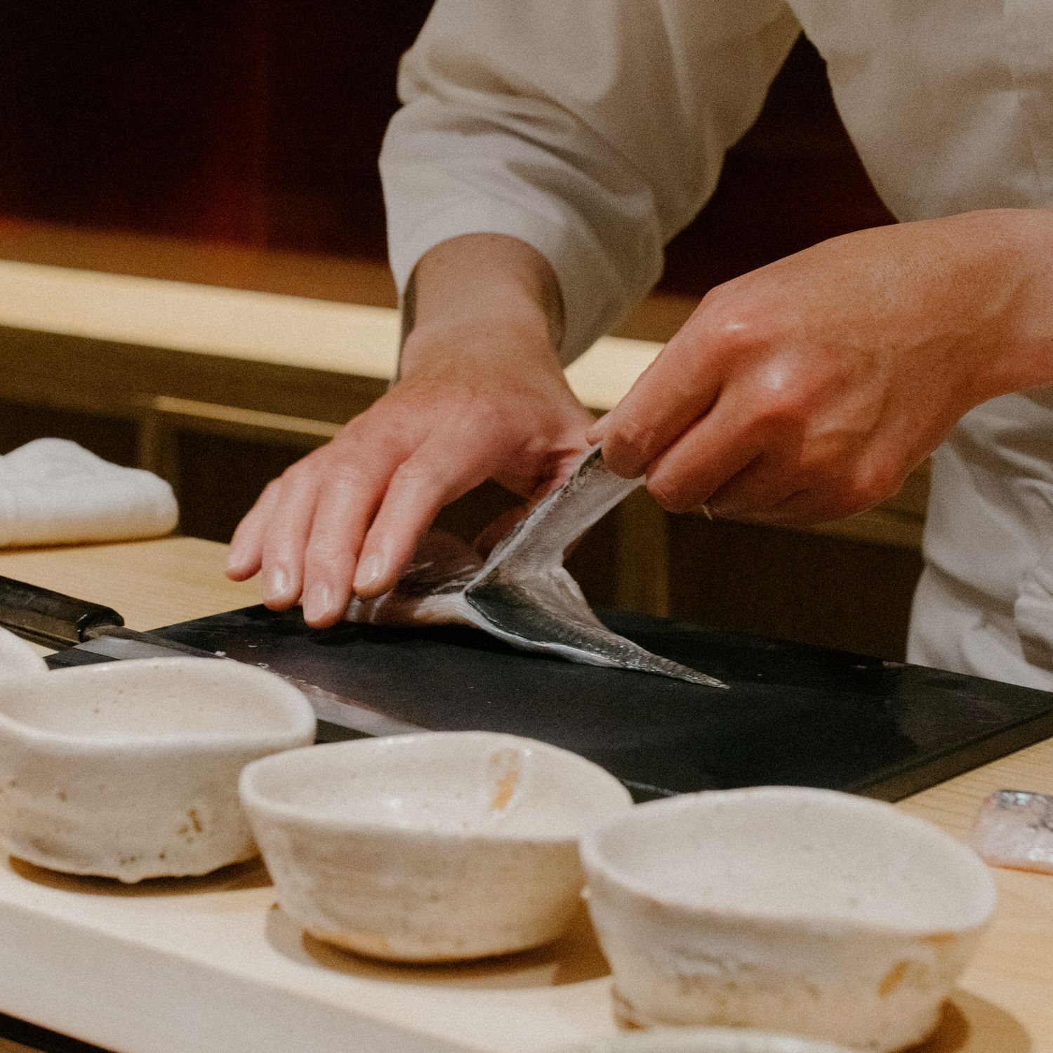 Close-up of hands slicing fish on a black cutting board with ceramic bowls nearby.