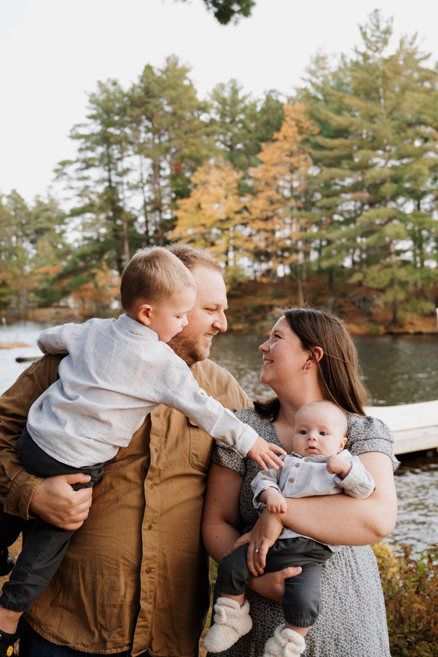 A family shares a tender moment together by a lake surrounded by autumn trees during a fall photoshoot.