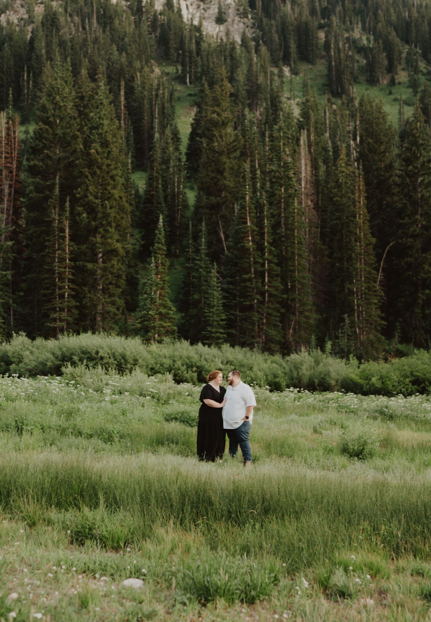 Couple walking together in a scenic mountain meadow surrounded by tall evergreen trees.