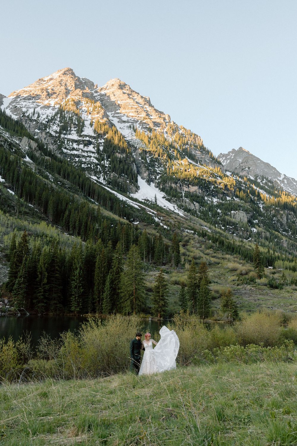 A couple in wedding attire stands in a meadow beneath snow-capped mountain peaks and evergreen forest at sunset.