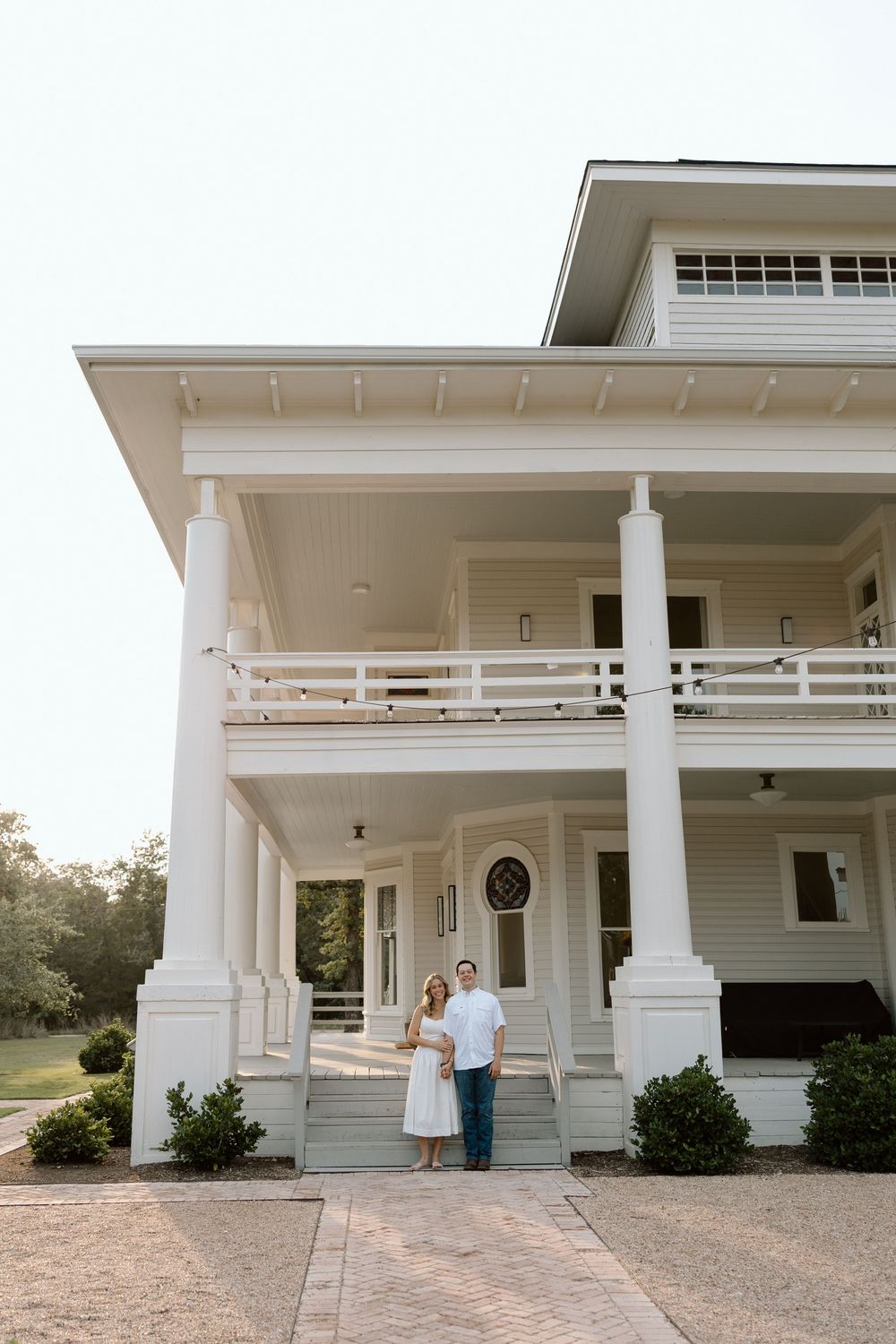 A southern mansion with white columns and wraparound porches features a couple standing on its front steps at sunset.