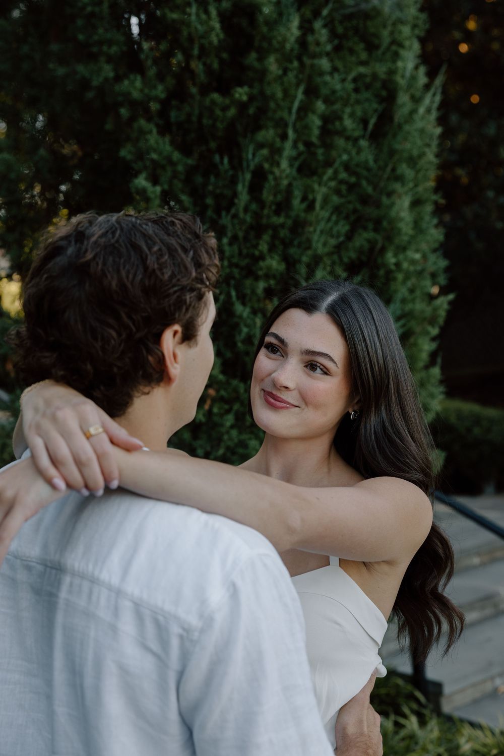 A romantic couple embraces outdoors against a backdrop of green cypress trees at dusk.