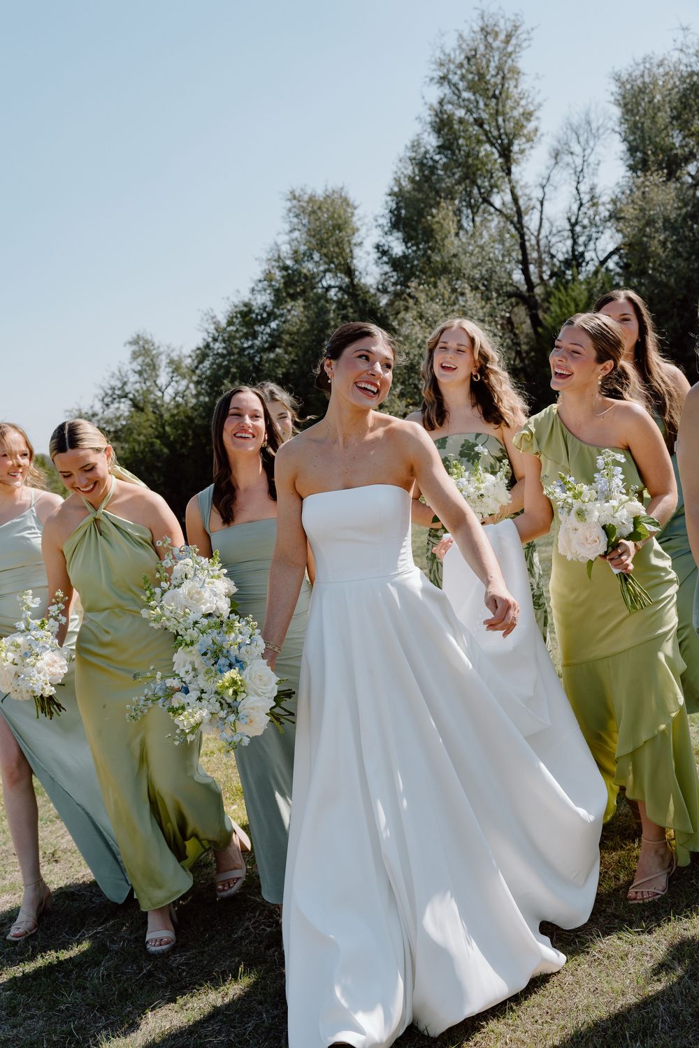 A bridal party wearing sage green dresses and holding white floral bouquets walks together outdoors on a sunny day.