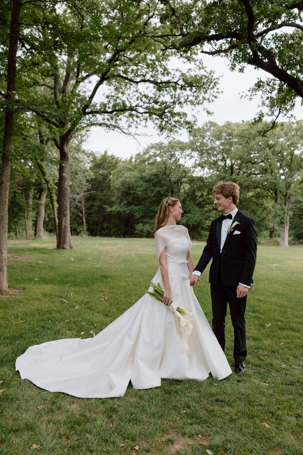 Bride and groom holding hands in a lush green park, bride wearing a white ball gown with train, groom in classic black tuxedo.